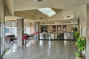 A hotel reception area with a reception desk, a cash register, and a sign that reads 'RECEPTION CASHIER.' There are potted plants and a vase with flowers on the reception desk.