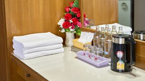 A hotel room with a wooden wardrobe, a white countertop, a mirror, a vase with red flowers, a stack of white towels, a coffee maker, and a trash bin.