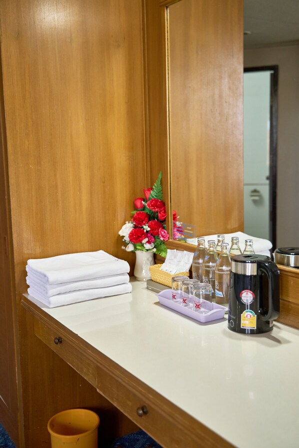 Habitación de hotel con un armario de madera, una encimera blanca, un espejo, un florero con flores rojas, una pila de toallas blancas, una cafetera y un cubo de basura.