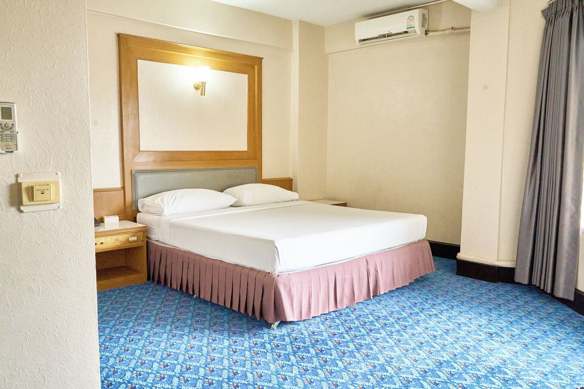 Een hotelkamer met een bed, een nachtkastje, een ingebouwde airconditioner, een spiegel en een kamerbreed tapijt.