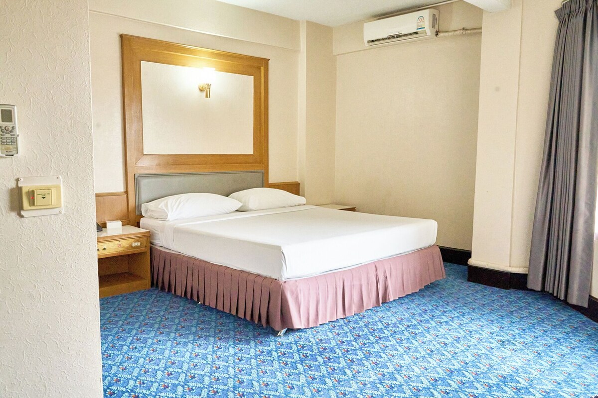 Habitación de hotel con cama, mesita de noche, aire acondicionado de pared, espejo y alfombra estampada.