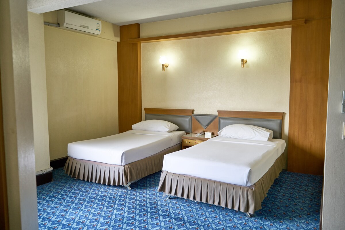 Habitación de hotel con dos camas, una mesita de noche, un equipo de aire acondicionado y una alfombra estampada.
