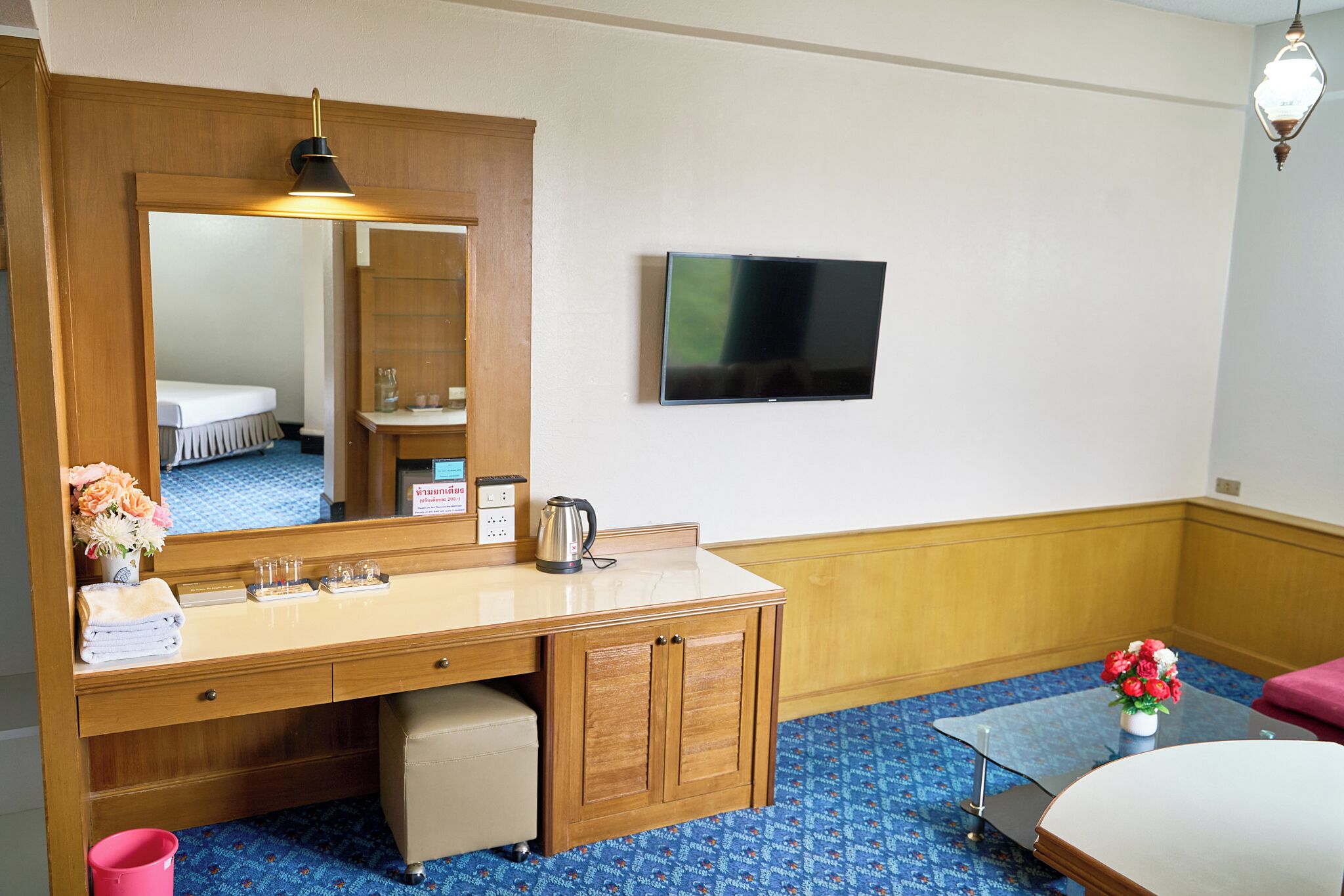 Habitación de hotel con tocador de madera, televisor de pantalla plana, espejo, pequeña mesa con un jarrón de flores y cama con alfombra de diseño azul.