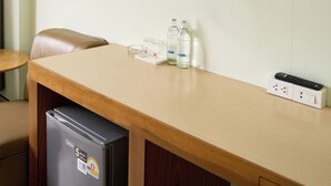 A hotel room with a flat-screen TV mounted on the wall, a wooden desk with a refrigerator, and a remote control on the desk.