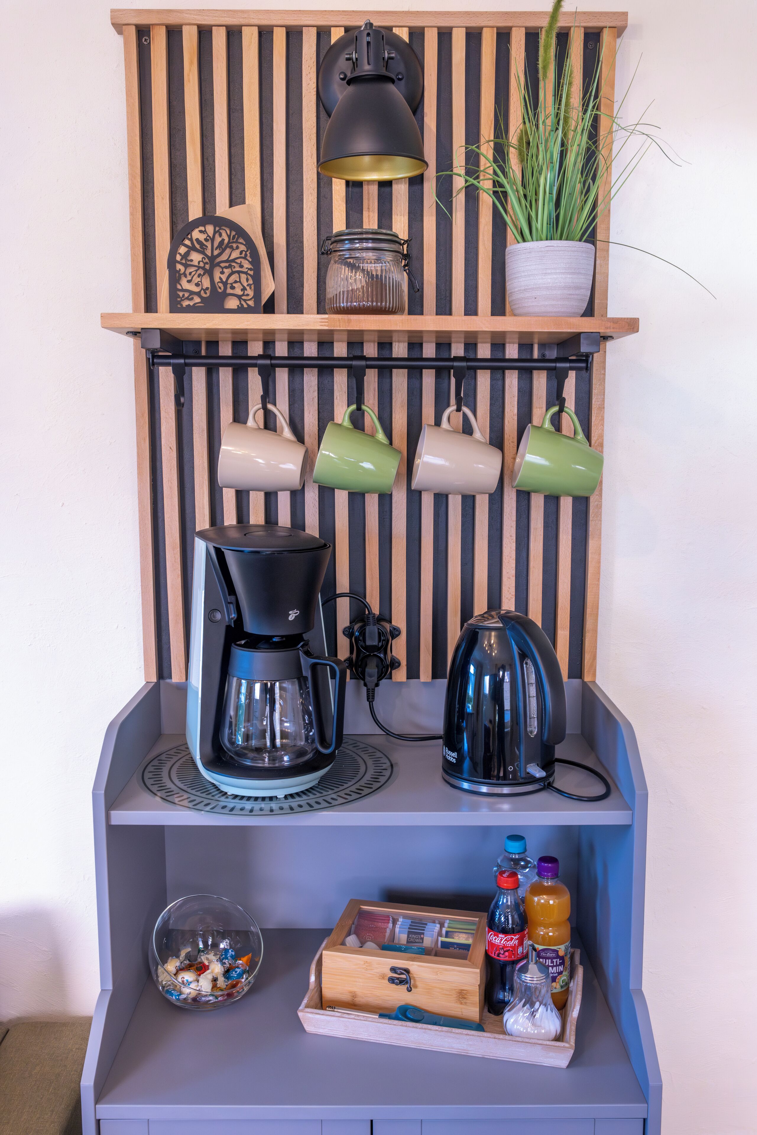 A kitchenette with a coffee maker, kettle, and mugs on a shelf, a potted plant, and a small cabinet with snacks and drinks.