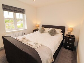 A neatly made bed with pillows, a bedside table with a lamp, a window with curtains, and a radiator under the window. - 11 The Forge (Brampton)