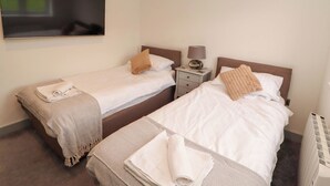 Two single beds with white linens, a small bedside table with a lamp, and a wall-mounted TV. - 11 The Forge (Brampton)