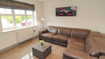 Uma sala de estar com um sofá de couro marrom, uma pequena mesa lateral com um vaso, um radiador, uma janela com vista para um carro vermelho e um quadro de um carro de corrida na parede.