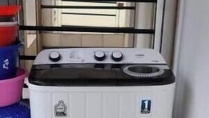 A compact, white washing machine in a corner with a tiled floor and a metal railing in the background. - Cozy 1-Bedroom Apartment in Westpark Heights (flyover)