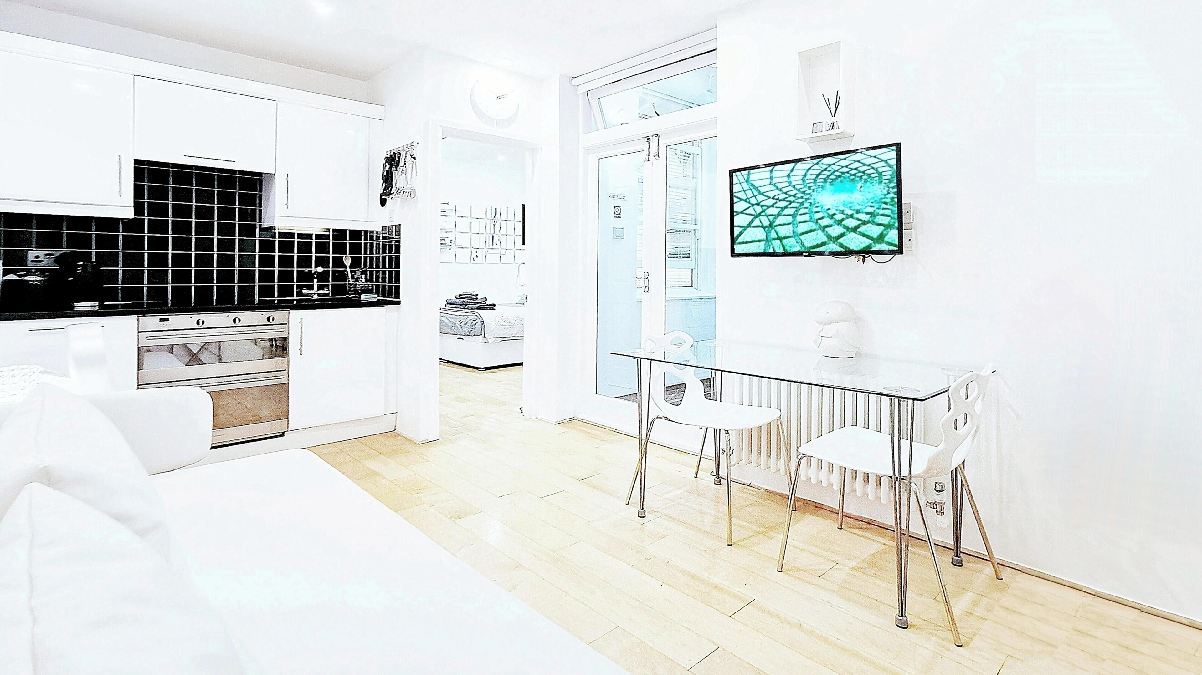 Une cuisine moderne avec des meubles blancs, un dosseret en carreaux noirs et un coin repas comprenant une table en verre et des chaises blanches.