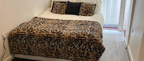 A bedroom with a bed, a leopard print bedspread, a black rug, and a window with sheer curtains.