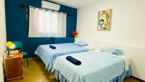 Una habitación con dos camas, un escritorio, una silla, un ventanal con cortinas y un aire acondicionado de pared.