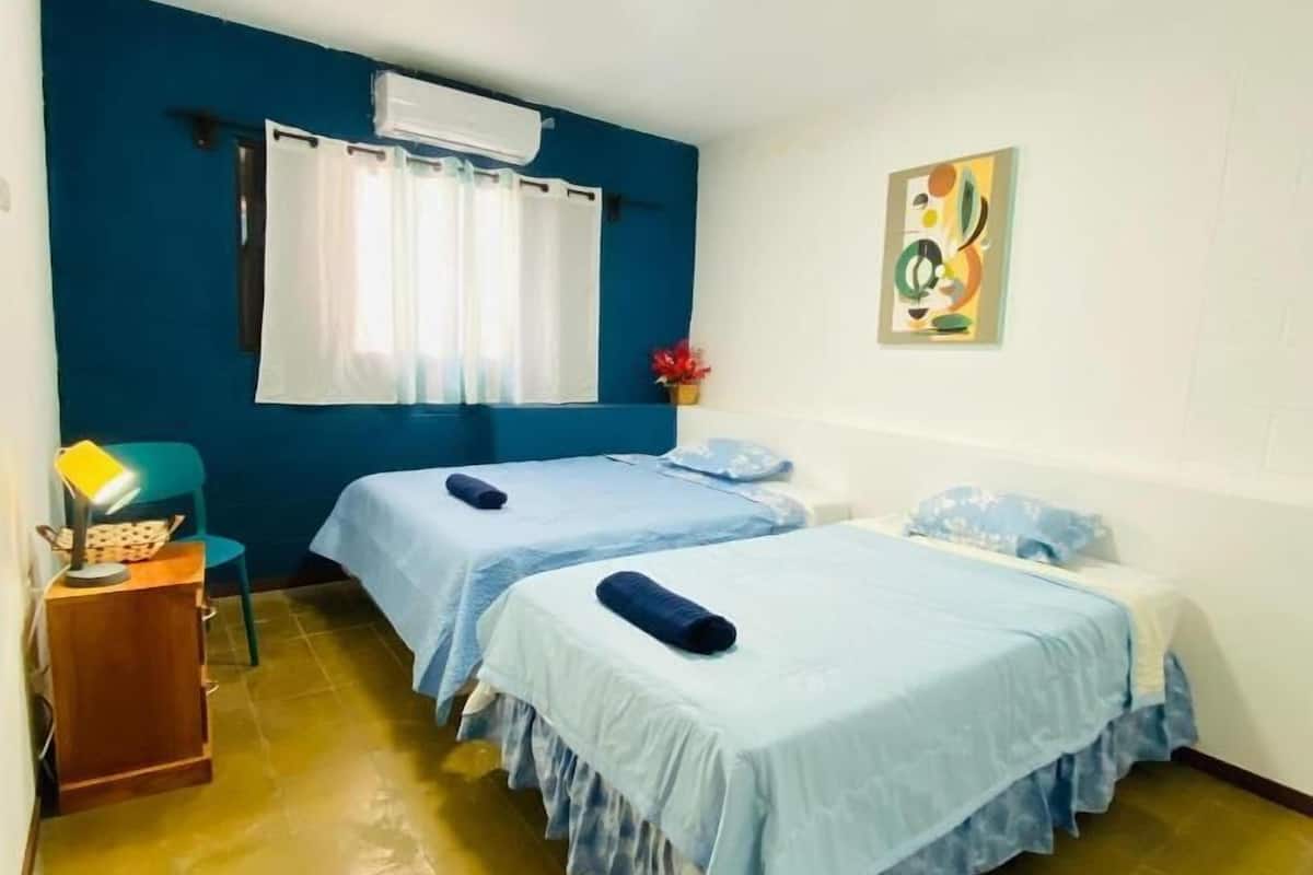Una habitación con dos camas, un escritorio, una silla, un ventanal con cortinas y un aire acondicionado de pared.