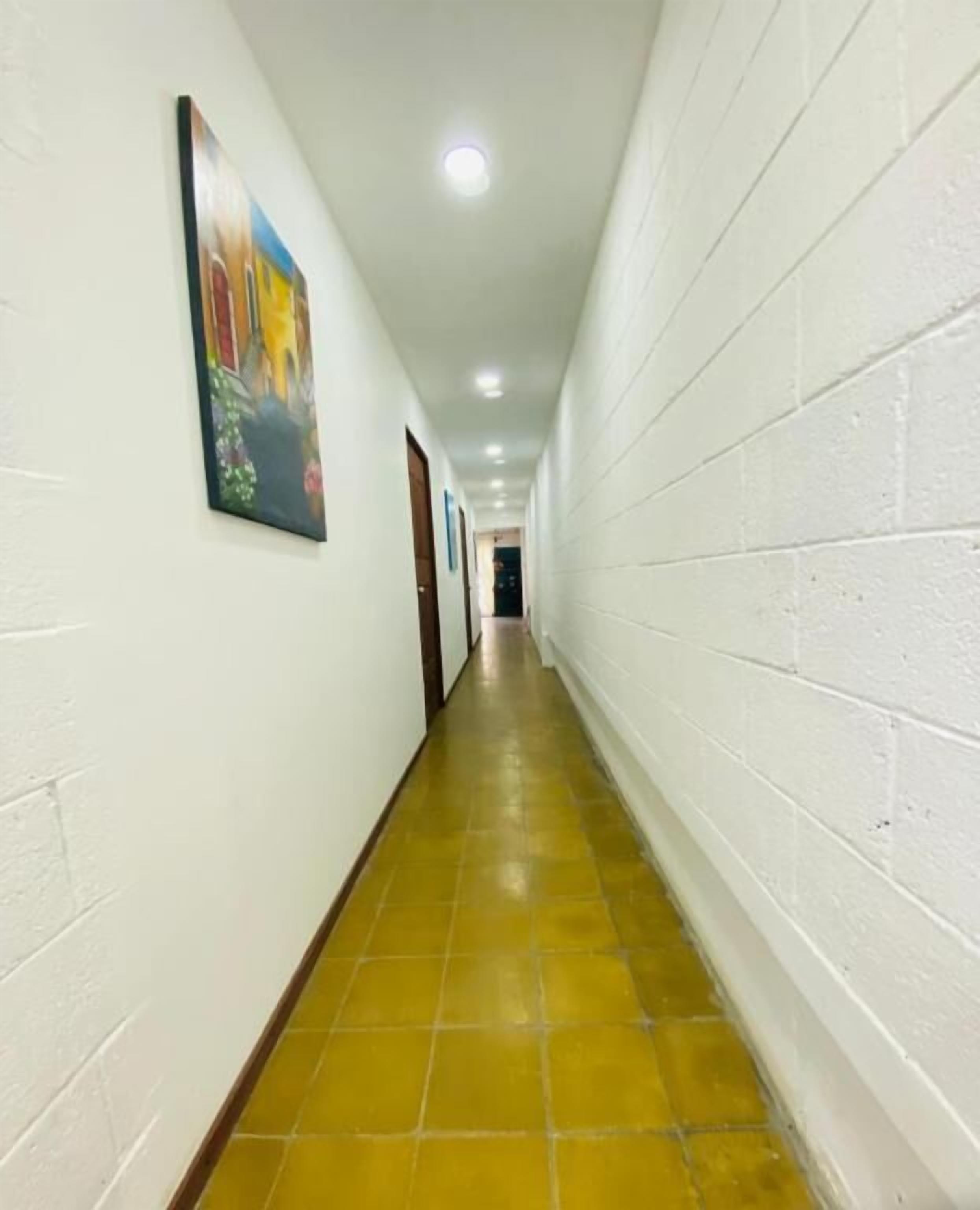 A long, narrow hallway with white brick walls, yellow-tiled flooring, and a ceiling with recessed lighting.