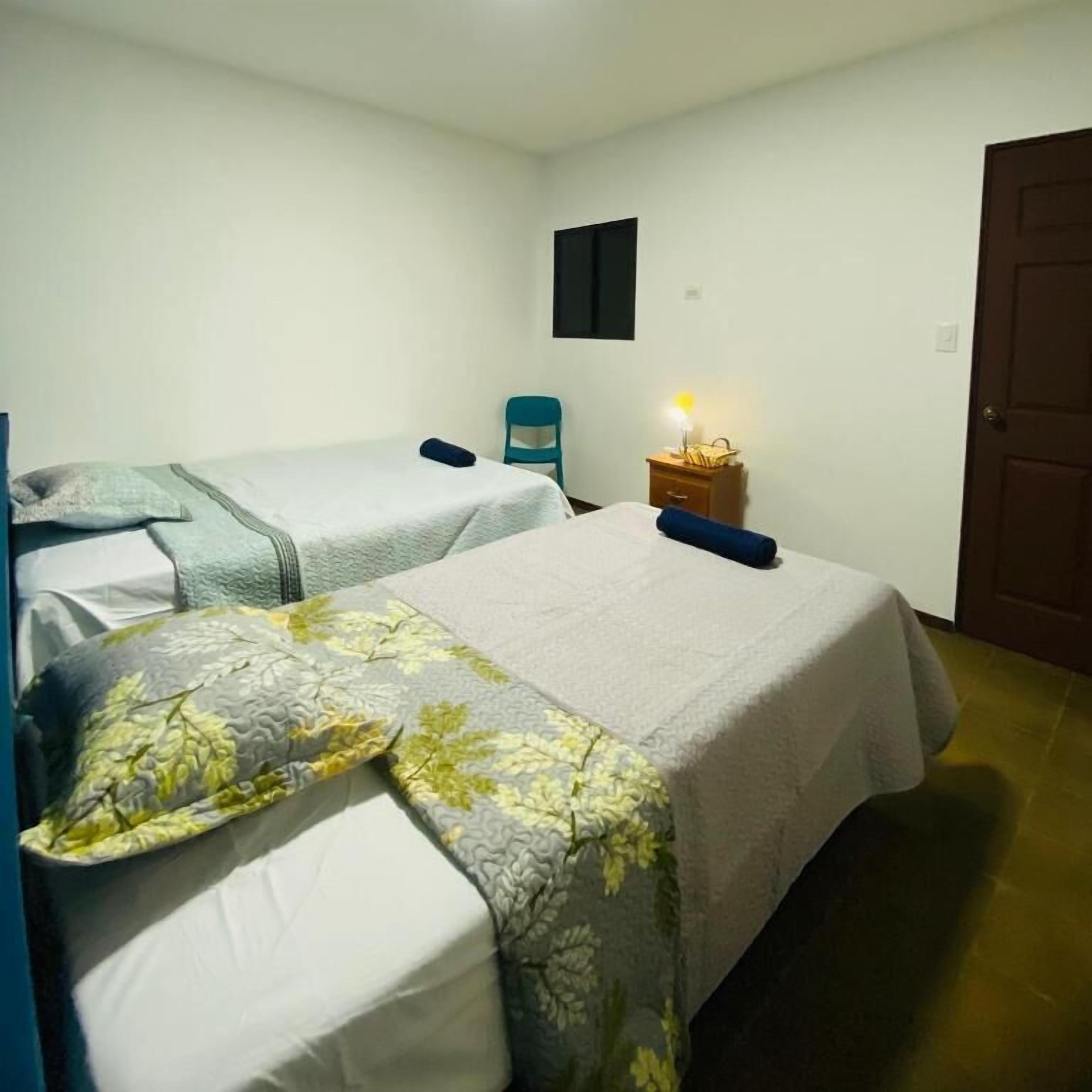 A room with two beds, a chair, a nightstand with a lamp, and a door.