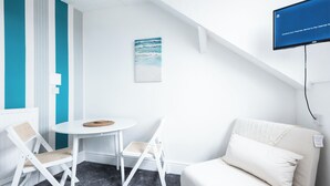 Ein Zimmer mit einem weißen Sofa, einem kleinen runden Tisch mit einer gewebten Unterlage, einem weißen Klappstuhl, einem wandmontierten Flachbildfernseher und einem gerahmten Bild vom Meer.