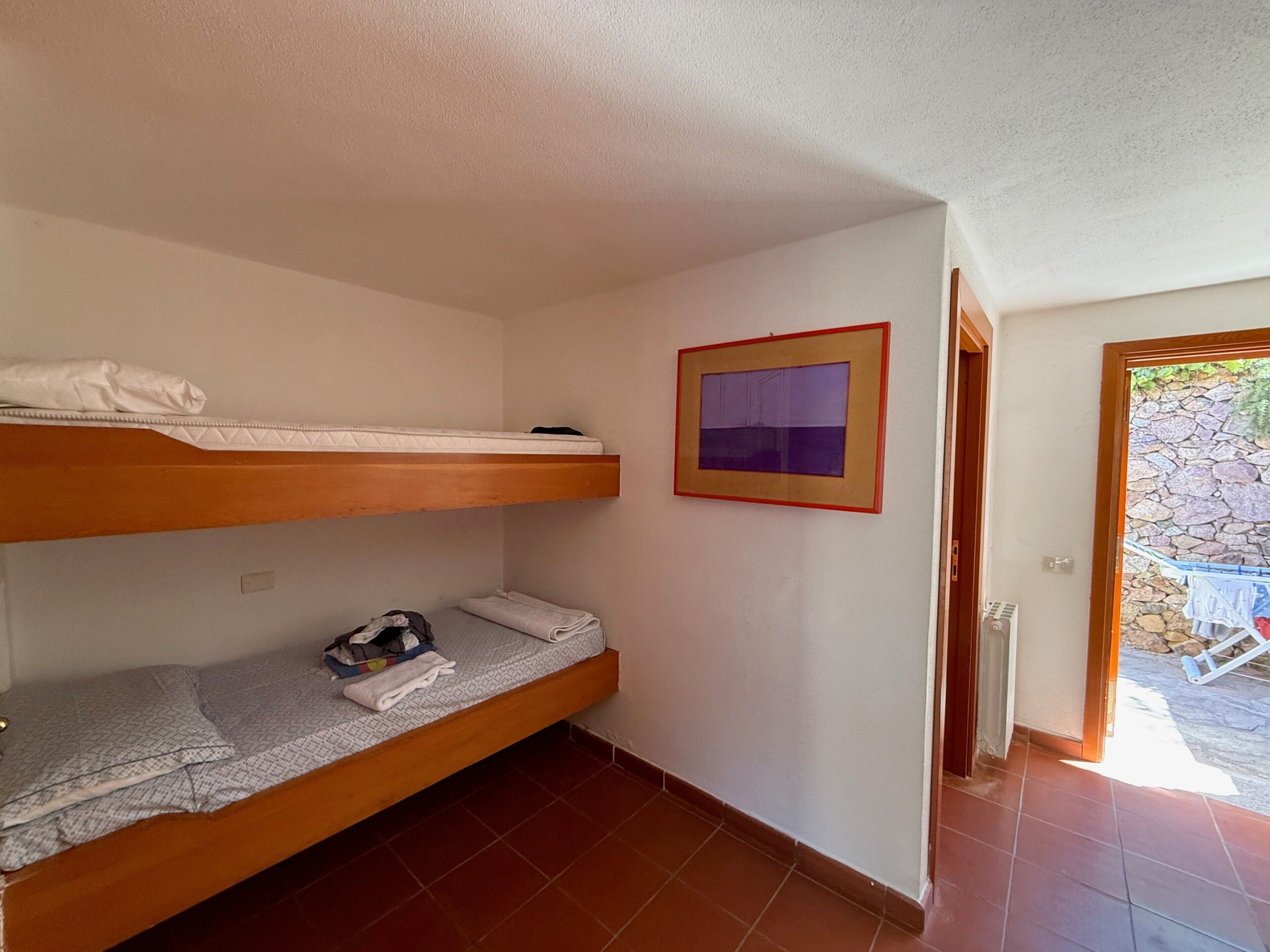 A room with two bunk beds, a framed picture on the wall, and a door leading to an outdoor area with a stone wall.