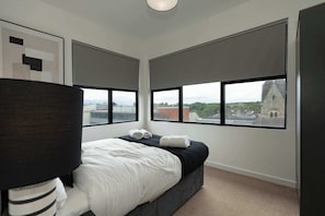 A modern hotel room with a large bed, two windows with blinds, a view of a cityscape, and a ceiling light. - Modern one-bedroom residence in Southampton Centre (Southampton)