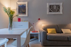 Een moderne woonkamer met een bank, een eettafel, een vaas met bloemen, een lijst met een afbeelding en een lamp.