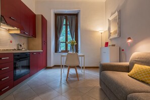 Een compacte leefruimte met een kitchenette, een zithoek met een tafel en stoel, een bank, een raam met gordijnen en een lamp.