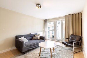 A modern living room with a sofa, a leather chair, a round coffee table, and a large window with curtains. - Belgravia Living Modern 2-bed Flat Near Victoria (London)
