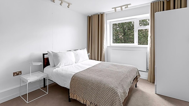 A bedroom with a bed, bedside table, lamp, window with curtains, and a wardrobe. - Belgravia Living 2-bed, 2-bath Fourth Floor Apartment (London)