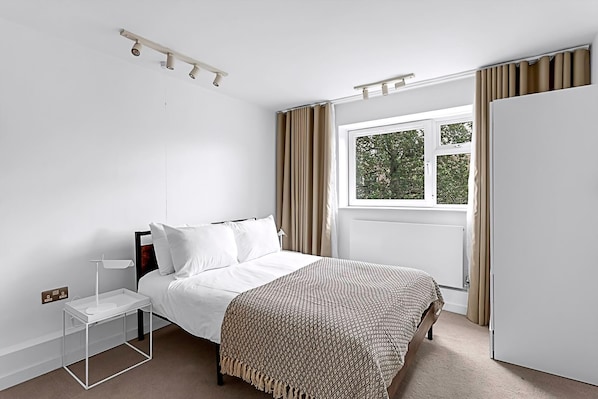 A bedroom with a bed, bedside table, lamp, window with curtains, and a wardrobe. - Belgravia Living 2-bed, 2-bath Fourth Floor Apartment (London)