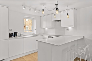 A modern kitchen with white cabinetry, a large island, pendant lighting, and a view of a brick building outside. - Belgravia Living 2-bed, 2-bath Fourth Floor Apartment (London)