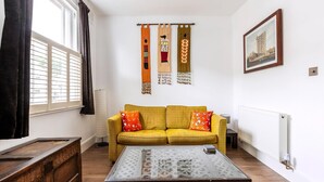 A room with a yellow sofa, a wooden coffee table, a framed picture on the wall, and a window with curtains. - Charming Golborne Escape (London)