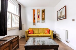 Ein Zimmer mit einem gelben Sofa, einem Holztisch, einem gerahmten Bild an der Wand und einem Fenster mit Vorhängen.