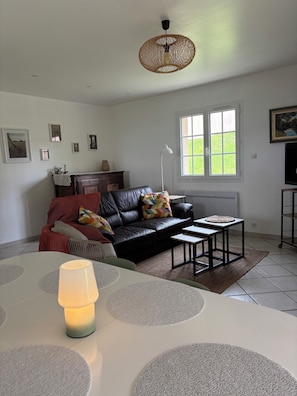 Un salon avec un canapé en cuir noir, une table ronde avec une lampe, un luminaire suspendu en osier et une fenêtre donnant sur un paysage verdoyant.