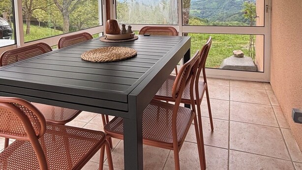 Un espace extérieur aménagé pour les repas, avec une table et des chaises, de grandes fenêtres offrant une vue sur les arbres et les collines, et un plafond en bois.