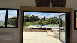 Une pièce dotée d’une grande porte-fenêtre coulissante donnant sur un patio extérieur avec une piscine, des chaises longues et une vue sur un paysage rural.