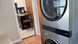 A laundry room with two stacked front-loading washing machines, a wooden chair, and a kitchen area visible through an open door. - 3-bedroom Cozy Home Close to Santa Claus, IN and Holiday World (Ferdinand)