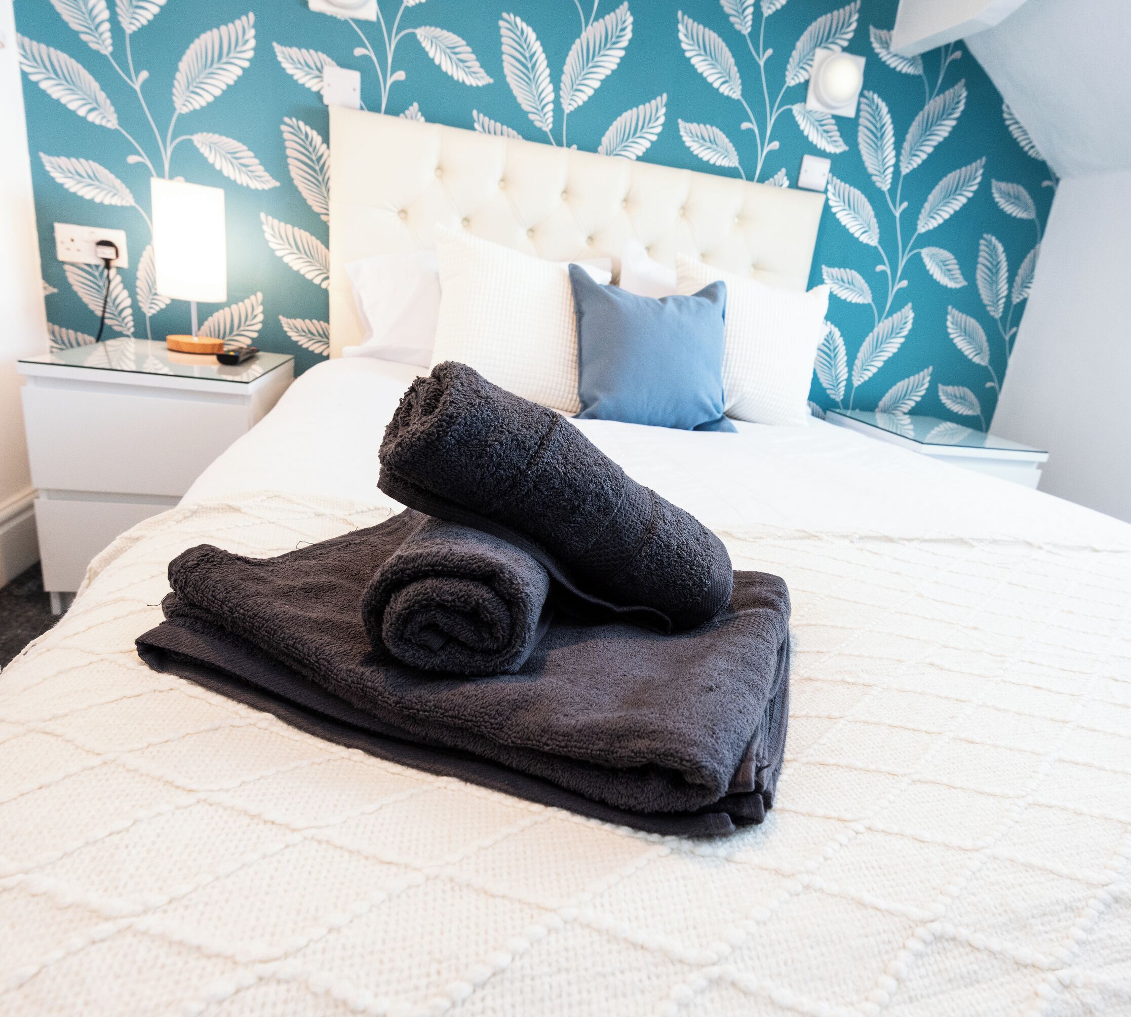 Een net opgemaakt bed met een wit hoofdbord, een opgerolde donkere handdoek en een witte geborduurde deken.