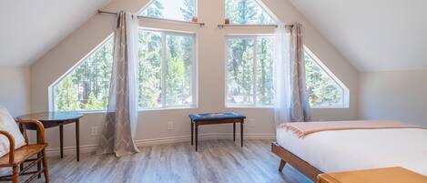 A bedroom with a sloped ceiling, large windows, a bed, a chair, and a small table.