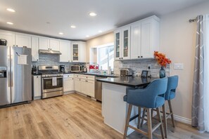 A modern kitchen with white cabinets, stainless steel appliances, a wooden floor, and a bar area with blue chairs. - Lakeview Cabin w/Walk to Lake + Slopes + Dog (Big Bear Lake)