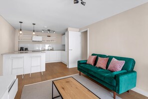 Eine moderne Küche mit weißen Schränken, einer Bar und einem petrolfarbenen Sofa mit rosafarbenen Kissen.