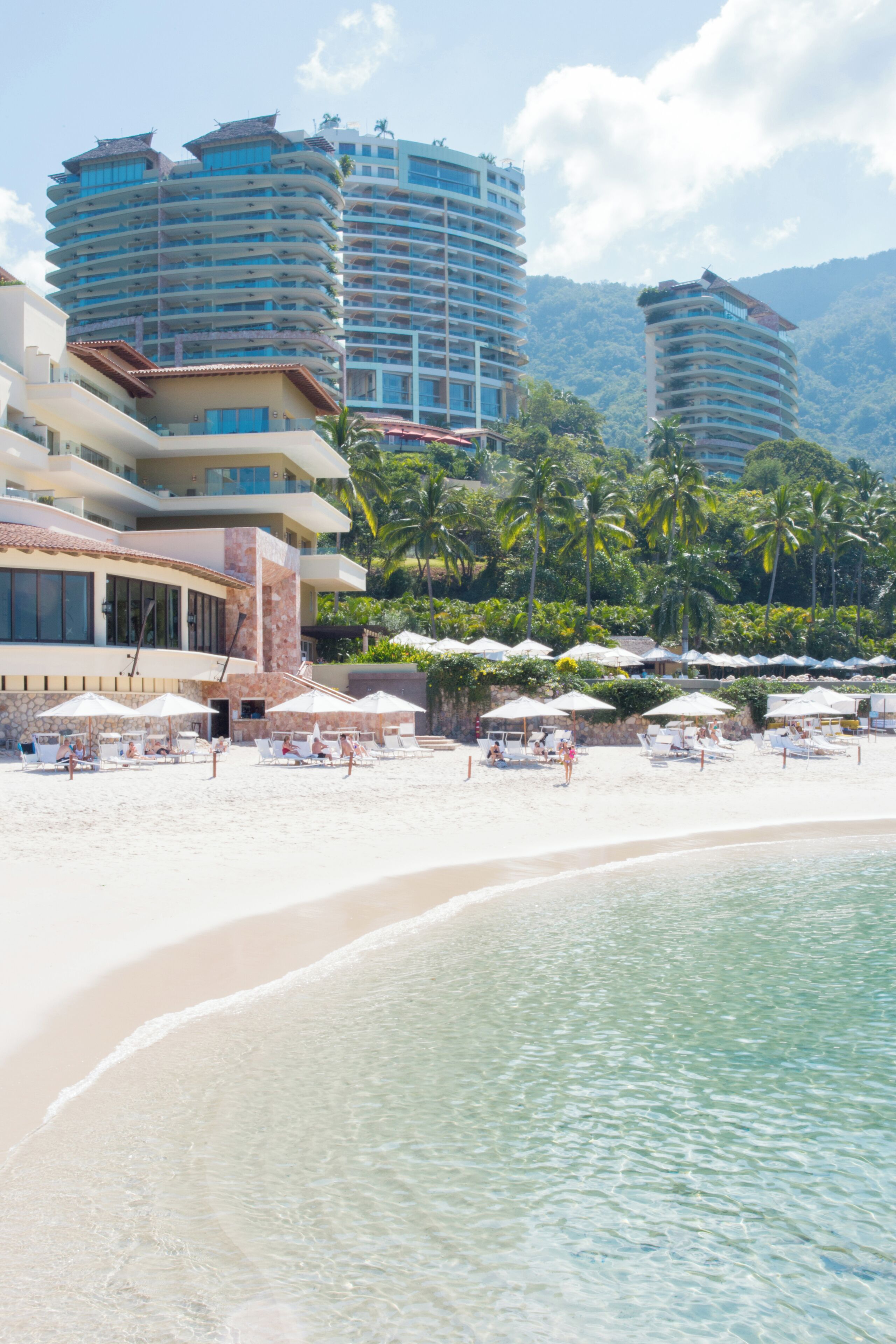 Un hôtel situé en bord de mer, doté de nombreux balcons, d’une plage de sable et de palmiers.