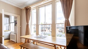 A room with a wooden dining table, a bench, a TV, and large windows with curtains. - Super Chic 1-bedroom Urban Hideaway (Twickenham)