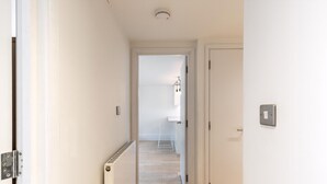 A narrow hallway with a radiator, a door with a light switch, and a view into another room with a radiator and a hanging light fixture. - Belgravia Living 2-bed, 2-bath Home With Balcony (London)