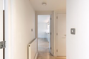 A narrow hallway with a radiator, a door with a light switch, and a view into another room with a radiator and a hanging light fixture. - Belgravia Living 2-bed, 2-bath Home With Balcony (London)