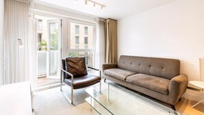 A modern living room with a sofa, a leather chair, a glass coffee table, and a view of a building through a sliding glass door. - Belgravia Living 2-bed, 2-bath Home With Balcony (London)