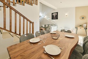 A dining area with a wooden table set for four, a staircase, and a living room in the background. - Villa Mascareignas · 3BR · 2 baths · 8 guests (Le Tampon)