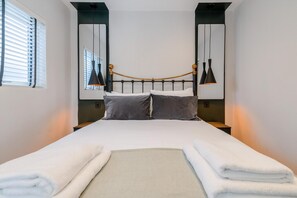 A neatly made bed with a headboard, two bedside lamps, and a window with blinds. - The Balham Hideaway (London)