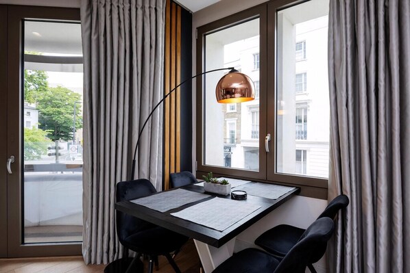 Ein modernes Zimmer mit Esstisch, Stühlen, einer Pendelleuchte, einem Fenster mit Vorhängen und Blick auf Gebäude.