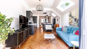 A modern interior with a kitchen, dining area, and living room, featuring a herringbone wood floor, a blue sofa, and a large window. - Stylish Family Home With Garden in Peckham Rye (London)