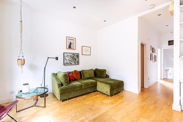 Ein Wohnzimmer mit einem grünen Ecksofa, einem Glastisch, einer Stehlampe und gerahmten Kunstwerken an der Wand.