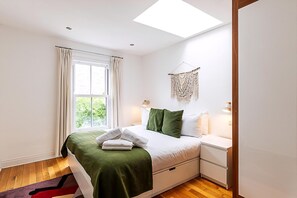 Ein Schlafzimmer mit Bett, Nachttischen, einem Fenster mit Vorhängen, einem Dachfenster und einem Wandbehang aus Makramee.