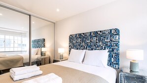 Habitación de hotel con una cama grande, una mesita de noche con lámpara, un espejo y una ventana con vistas a edificios.